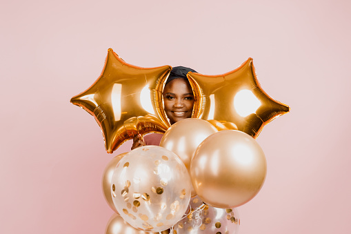Black girl between golden ballons smiles and have a party isolated on blank pink background for advert . African woman celebrate holiday. Happy emotions of muslim young woman