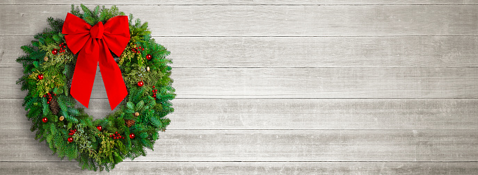 An evergreen Christmas wreath with red ornaments hangs from a whitewashed wall.