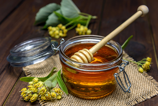 Jar with linden honey with stick for honey and fresh linden flowers on a wooden table.