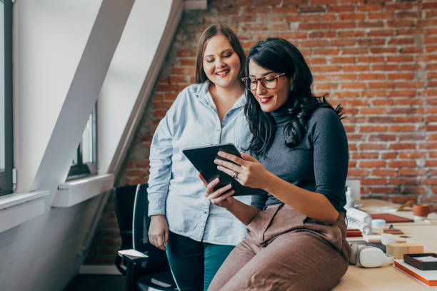 デジタルタブレット上の何かを見てオフィスで一緒に働く2人の幸せな若いビジネスウーマン - digital tablet businesswoman young women watching ストックフォトと画像