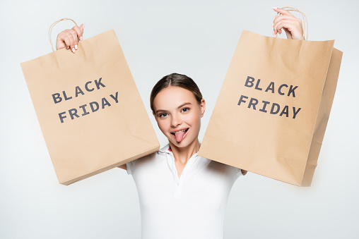 young woman sticking out tongue and holding shopping bags with black friday lettering isolated on white