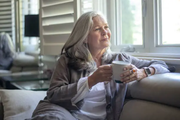 A senior woman with long white hair enjoys a coffee whilst sitting on a sofa. She is looking out the window and pondering about her retirement.