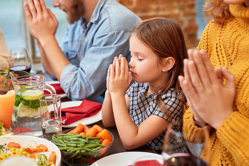 Little girl sitting near mother, praying before eating, holding hands on served dinner table with homemade food. United christian family spending time together, celebrating traditional easter