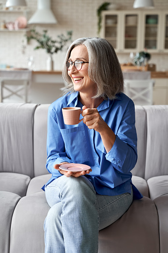 Happy beautiful relaxed mature older adult grey-haired woman drinking coffee relaxing on sofa at home. Smiling stylish middle aged 60s lady enjoying resting sitting on couch in living room, vertical.