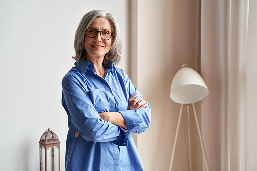 Smiling confident mature older woman standing arms crossed indoors looking at camera. Stylish classy middle aged senior 60s grey-haired lady wearing blue elegant shirt and glasses indoors, portrait.