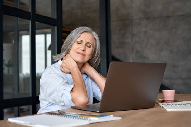 疲れたストレスの古い成熟したビジネスウーマンは、テーブルに座ってオフィスで働く線維筋痛症の首の痛みに苦しんでいます。過労の先輩中年女性は、座りっぱなしの仕事から痛みを感じ� - spasm ストックフォトと画像