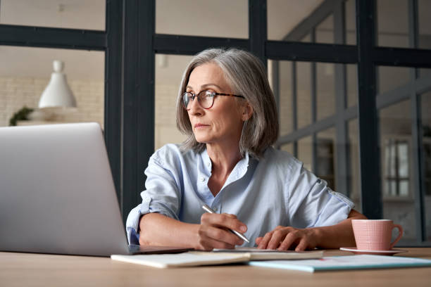 femme adulte plus âgée sérieuse regardant le webinaire de formation sur l’ordinateur portatif fonctionnant de la maison ou dans le bureau. femme d’affaires d’âge moyen des années 60 prenant des notes tout en utilisant la technologie d’ordinate - adulte dâge mûr photos et images de collection