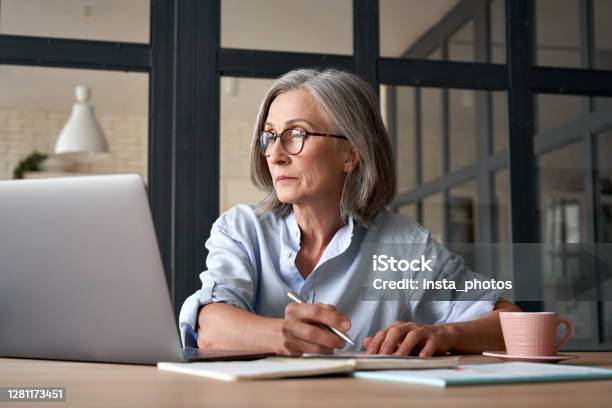 Serious Mature Older Adult Woman Watching Training Webinar On Laptop Working From Home Or In Office 60s Middle Aged Businesswoman Taking Notes While Using Computer Technology Sitting At Table Stock Photo - Download Image Now