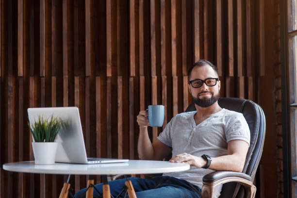 istock Young successful bearded businessman freelancer in office coworking. Happy man programmer developer in a white t shirt smiling and drinking coffee at the Desk with a laptop 1281184513