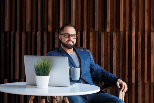 istock Young successful bearded businessman freelancer in office coworking. Happy man programmer developer in a white t shirt smiling and drinking coffee at the Desk with a laptop 1281184530