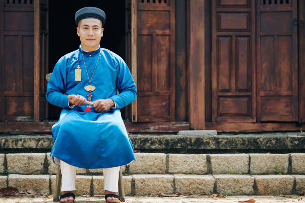 Man in ao dai dress Handsome young man in traditional ao dai dress sitting on stool in front of old temple ao dai stock pictures, royalty-free photos & images