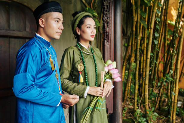 Serious young Vietnamese couple in blue and green ao dai dresses standing at entrance doors of old temple