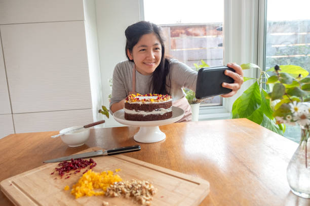 Side Hustle, Vegan Food Blogger Taking Selfie with Decorated Cake Eurasian young woman filming homemade vegan spice cake with walnuts and dried cranberries and apricots in kitchen on smartphone.  Vancouver, British Columbia, Canada. decorating a cake photos stock pictures, royalty-free photos & images