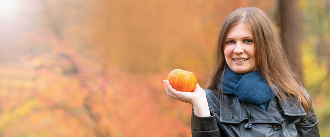 The girl and the pumpkin. A beautiful girl smiles in a leather jacket and scarf holding a small pumpkin in her hand against the background of an autumn forest. Halloween. Banner