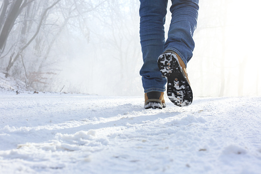 Man walking on a frosty road in a beautiful winter morning with sun and fog. Close-up to legs, snow on sole of boots.