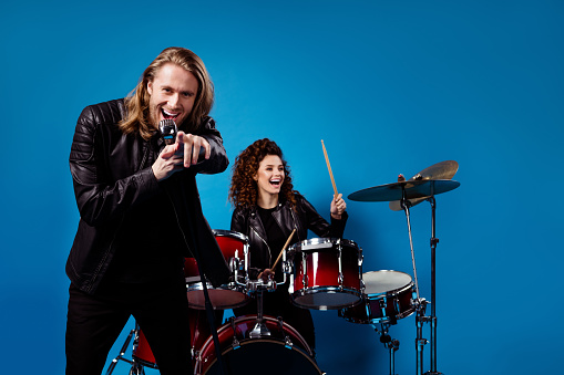 Portrait of funky famous rock band youth team two people man point index finger, sing song mic woman play drums enjoy sound garage studio event tour isolated over blue color background