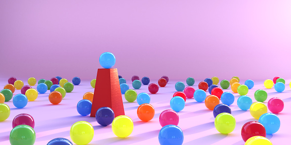A group of shiny, multi coloured spheres randomly arranged on a pink floor surrounding a single sphere placed higher up on a red plinth.