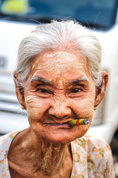 Old woman Woman over 80 years old. Smiling. He has a Myanmar roll cigarette in his mouth. Again on his face is the mixture, the traditional sun protection method of Myanmar.The woman is looking at the camera. Portrait shooting. Myanmar, Yangon center bazaar. 02/29/2020 smokers wrinkles stock pictures, royalty-free photos & images