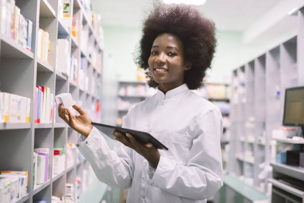 African American female pharmacist using digital tablet during inventory in pharmacy, holding pill box and smiling to camera African American female pharmacist using digital tablet during inventory in pharmacy, holding pill box and smiling to camera. pharmacy tech stock pictures, royalty-free photos & images