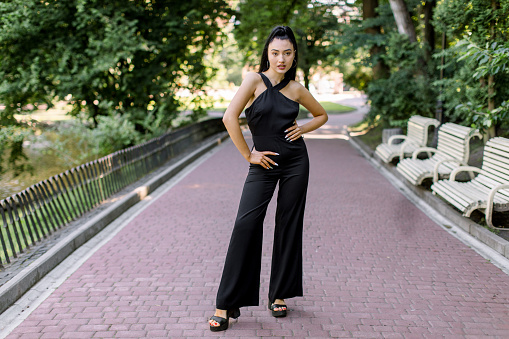 Full length portrait of young Asian woman model with ponytail hairstyle, smiling in urban park. Mixed raced girl wearing black trendy overalls and fashionable heels, posing on the park alley outdoors.