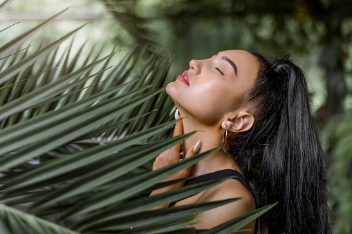 Horizontal beauty shot of pretty sensual Asian girl model, with long black ponytail hair, tounching neck with eyes closed while posing between exotic palm leaves. Natural beauty and people concept
