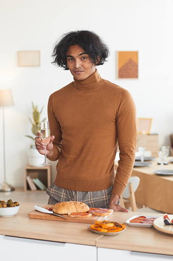 Vertical Waist up portrait of contemporary mixed-race man holding champagne glass and looking at camera while cooking for dinner party indoors