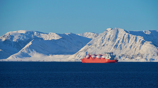 Gran buque portaaviones de GNL (gas natural licuado) pintado de rojo que se encuentra anclado en el océano ártico frente a la isla de S'rya con montañas cubiertas de nieve cerca de Hammerfest, Noruega en invierno. photo