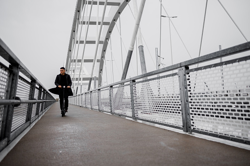 A young man in smart casual clothes riding an electric scooter over the bridge on a rainy Autumn day.