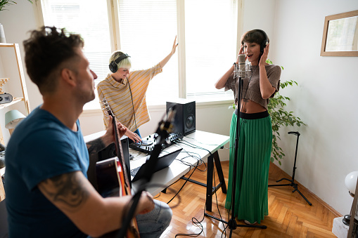 Young, Caucasian performance group made of female singer, female dj and male guitarist enjoying recording their new song in the home music recording studio.
