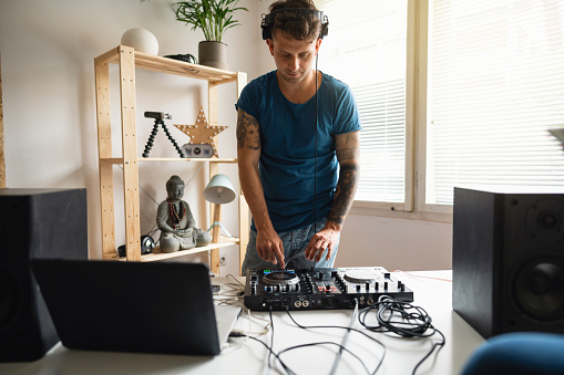 Young, Caucasian dj surrounded by audio equipment practicing mixing music at home. He uses dj control and laptop.