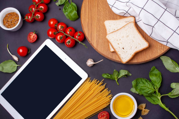 istock Cooking online. Tablet, cherry tomatoes, pasta, basil and bread on a black background 1281251490