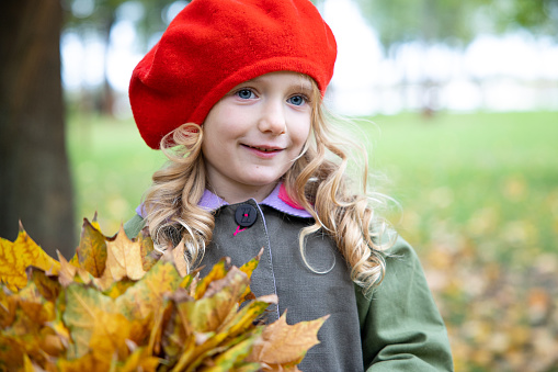 Portrait of a beautiful little preschool girl. A child walks with yellow maple leaves in the autumn park.