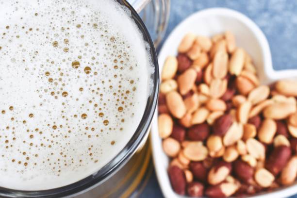 arachidi in ciotola a forma di cuore e birra su sfondo blu - healthy eating eating food and drink beer nuts foto e immagini stock