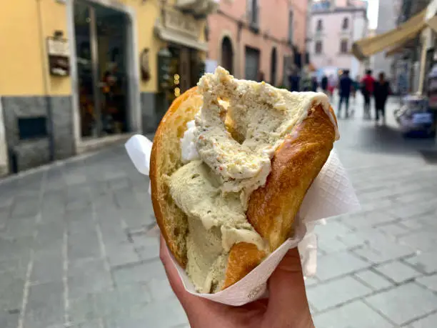 Traditional sicilian ice cream gelato in brioche bun in female hand close up on the background of Taormina street
