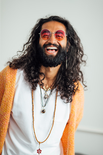 A portrait shot of a mid aged Pakistani man laughing and looking at the camera. He is wearing bohemian clothing and jewellery, sunglasses and has a large black beard and long curly hair.