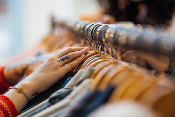 A close up shot of a woman's hand sifting through clothing hanging on a clothes rack. The woman has tattoos on her hands and is wearing a bracelet.