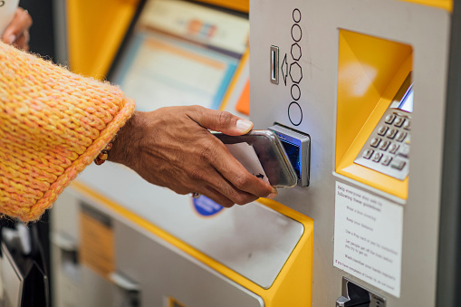 A shot of an unrecognisable man using his mobile phone to contactless pay for a train ticket on a ticket machine. The man is wearing wrist and finger jewellery and is holding his phone in one hand and a takeaway coffee cup in the other.