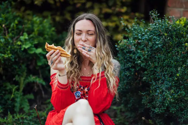 A shot of a young caucasian hipster woman sitting on a wall in an urban park. She is wearing casual bohemian clothing, accessories and is eating a toasted sandwich and giggling.