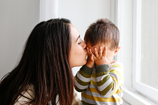 Portrait of young mother kissing her little son in forehead at home. Beautiful female with straight brunette hair playing with child at living room. Close up, copy space, background.