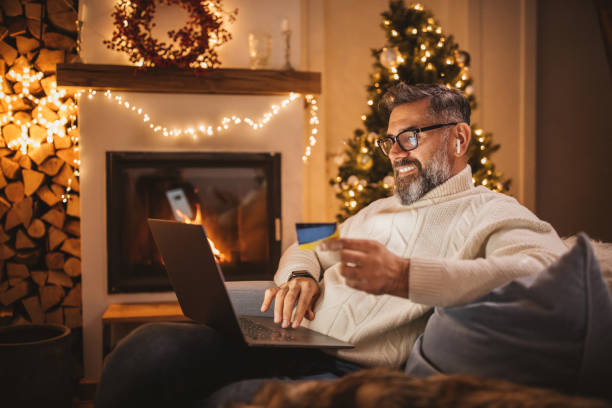 Mature man working from home Mature man sitting on couch at cottage for Christmas and using laptop. He is talking buying from home during pandemic isolation. one mature man only audio stock pictures, royalty-free photos & images