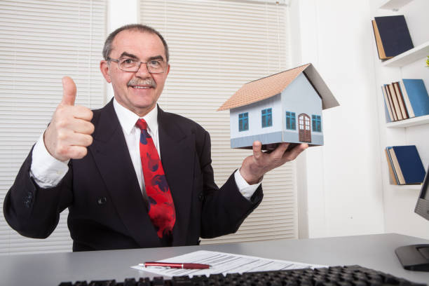 Happy man with beard wearing dark official style suit big paper key and pointing to camera with toothy smile, house purchase concept. Indoor studio shot isolated on blue background.