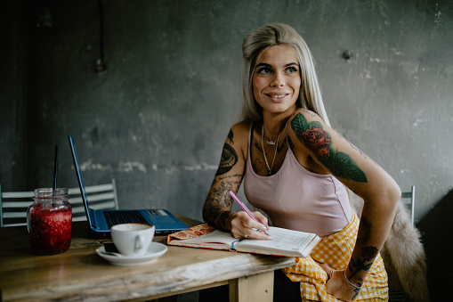 Beautiful young tattooed woman working, writing, and using a laptop in the coffee bar