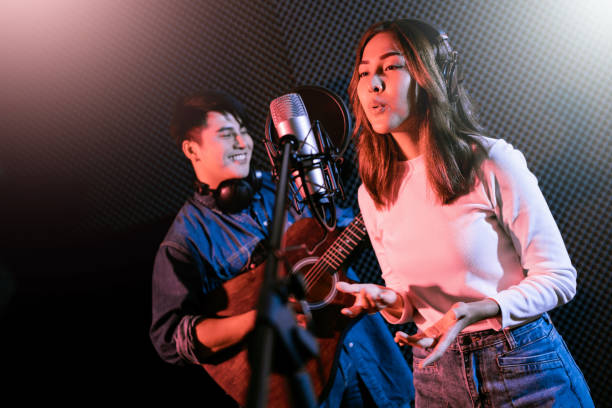 istock Pretty Asian female singer recording songs by using a studio microphone and pop shield on mic with male playing guitar in blue and red light. Performance and show in the music business. Duet session. 1281297286