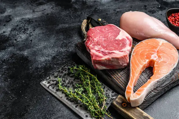 Photo of Set of raw meat steaks salmon, beef and chicken on a cutting board. Black background. Top view. Copy space