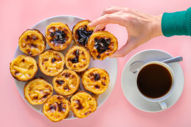 istock Hand Picking up a Pastel de Nata from a plate of Pasteis de Belem (aslo called Portuguese Egg Tarts or Pastel de Nata) on a pink background beside a cup of coffee, Food Flat Lay 1281302931