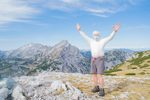 View of happy Senior  man with mask and hands up in ear  on Corona pandemical time on top of mountain over 2110 in Kamnik Savinja Alps, Slovenia.