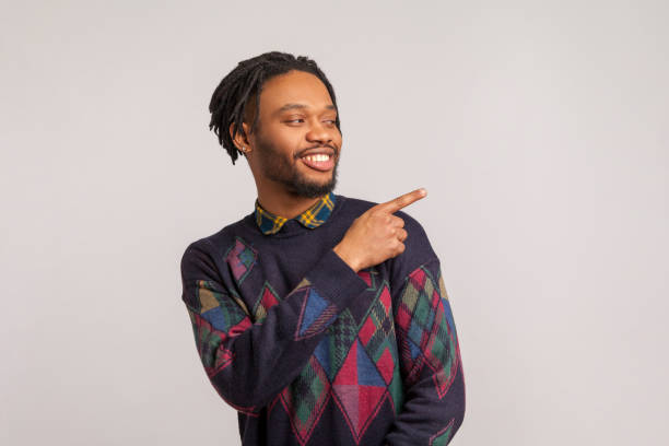 istock Cheerful mysterious african man with dreadlocks in stylish sweatshirt pointing finger away with toothy smile on face, empty space for your advertisement 1281323532