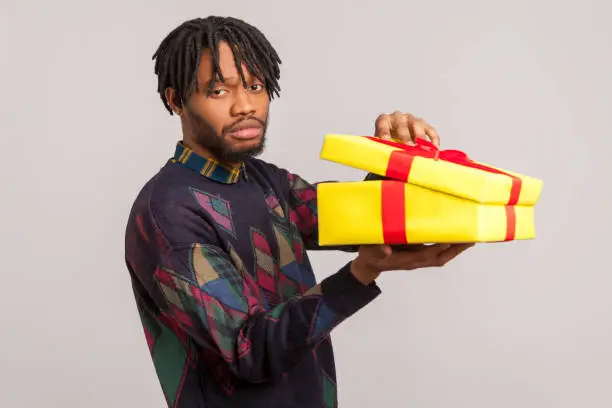 Unhappy disappointed african guy with dreadlocks holding unpacked gift box with frustrated dissatisfied face, upset with present. Indoor studio shot isolated on gray background