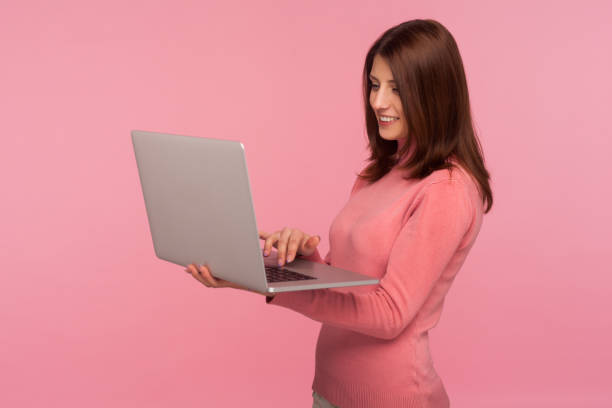 femme positive de sourire avec les cheveux bruns dans le chandail rose travaillant sur l’ordinateur portatif, travail indépendant, étude en ligne. - women computer home interior brown hair photos et images de collection