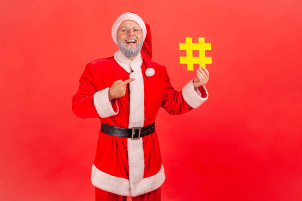 homme barbu gris heureux positif dans des lunettes et le costume de père noël pointant du doigt au symbole jaune de hashtag dans sa main, marquant des poteaux de vacances. - men suit holding human finger photos et images de collection